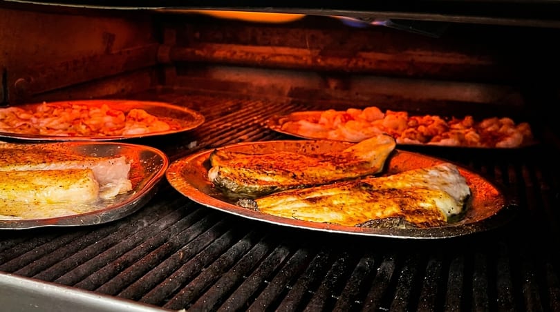 Fish fillets and shrimp cooking on metal trays inside a hot oven or wood-fired oven.