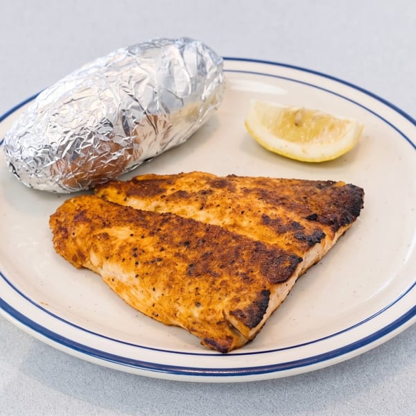 Blackened fish fillet with a foil-wrapped baked potato and lemon wedge on a white plate.