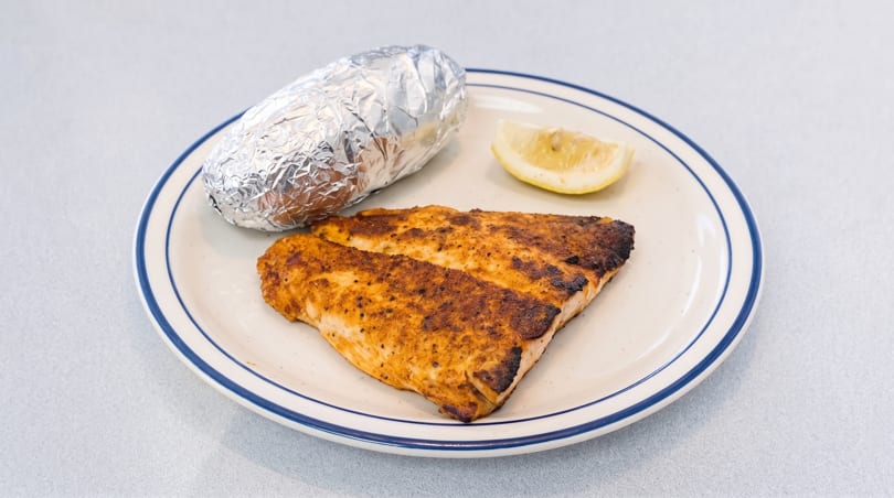 Blackened fish fillet with a foil-wrapped baked potato and lemon wedge on a white plate.